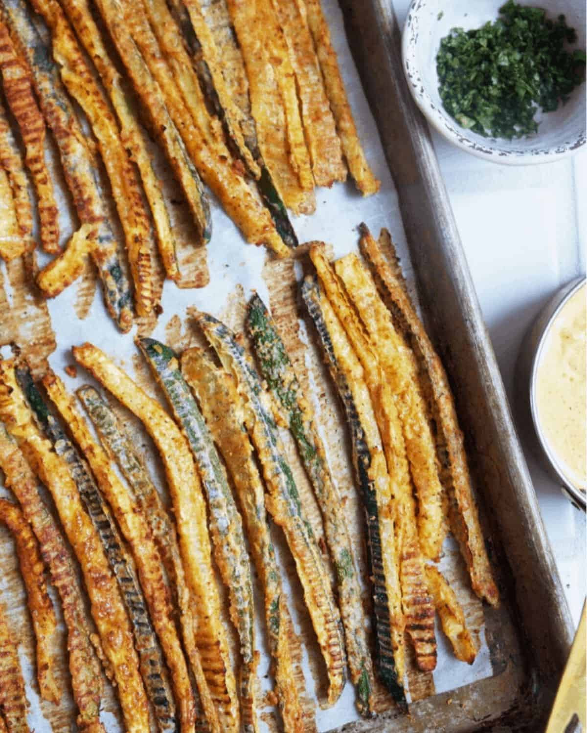 Baked zucchini fries arranged on a baking sheet in a single layer next to a bowl of herbs and container of dipping sauce. 