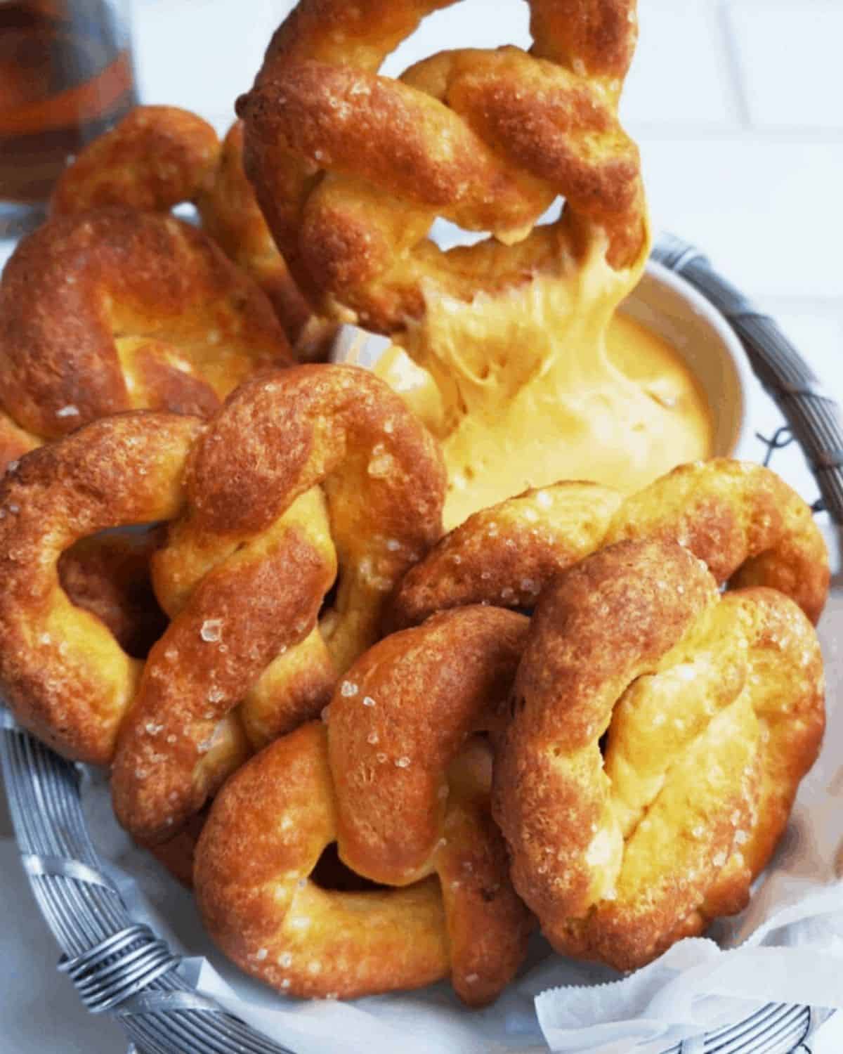 Six keto pretzels in a wire basket with one pretzel being dipped into cheese sauce