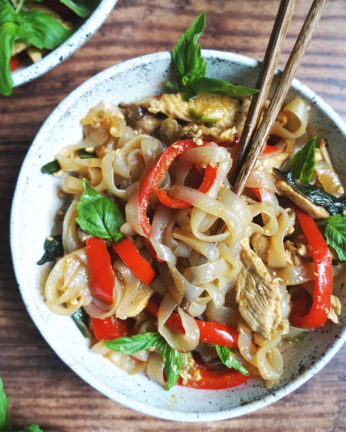 close up of a bowl of drunken noodles on a wooden table with chopsticks in it